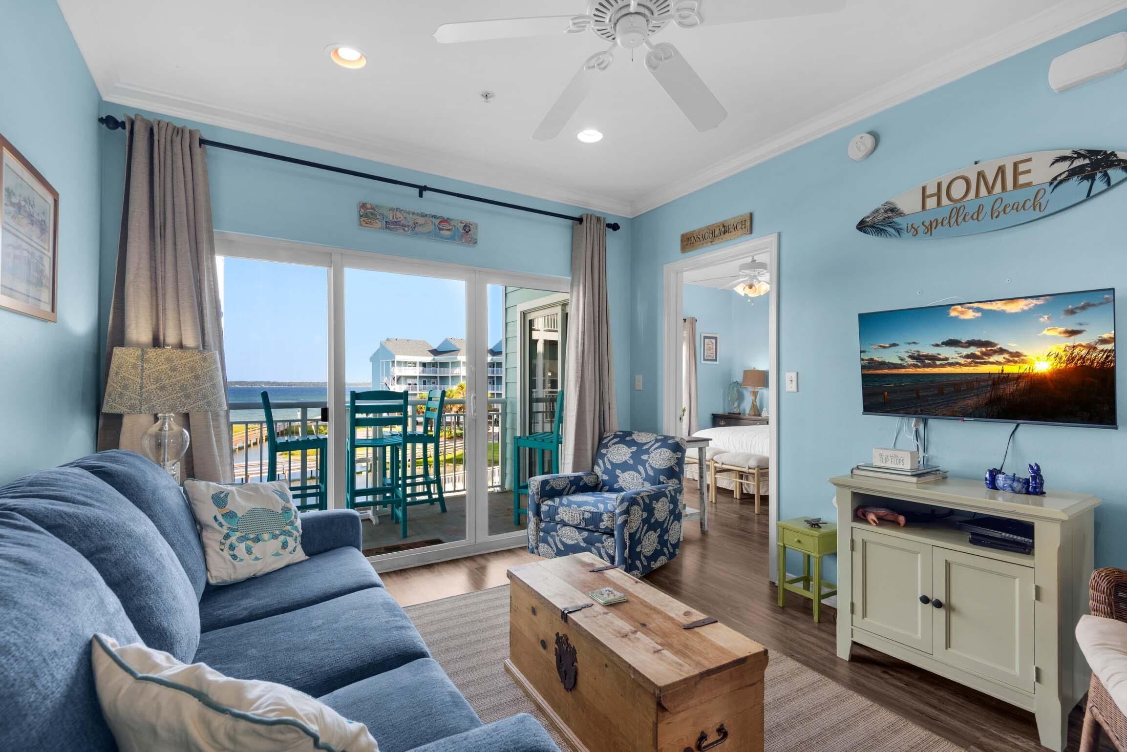 A living room with a blue sofa, a wooden coffee table, and a TV on a cabinet. There is a view of the ocean through large windows.