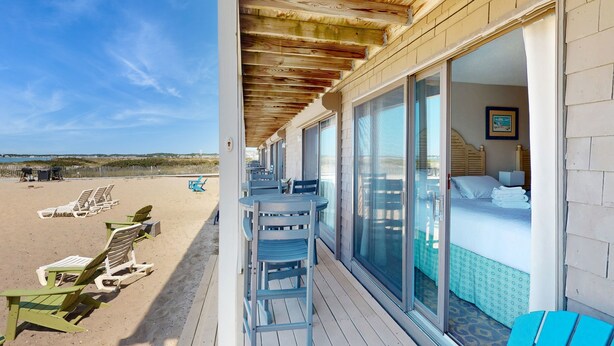 Une chambre d’hôtel située en bord de mer, avec une porte-fenêtre coulissante donnant sur la plage, un lit recouvert d’une courtepointe à motifs et une vue sur les chaises longues et les parasols.