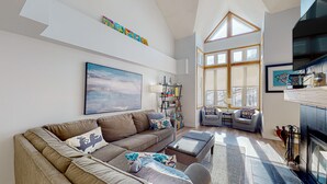 A modern living room with a sectional sofa, a coffee table, and a fireplace. There is a bookshelf and a window with a view of the outdoors. - Chateau Ridge (The Blue Mountains)