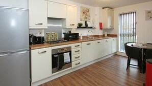 Una cocina moderna con armarios blancos, un refrigerador de acero inoxidable, un microondas y una estufa a gas. Hay una ventana con persianas y una pequeña zona de comedor con una mesa y sillas.