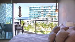 A hotel room with a large bed, a balcony with a table and chairs, and a view of a high-rise building and palm trees. - Spectacular Ocean View from 6th floor Beachfront Studio -Aria Ocean Flamingos NV (Nuevo Vallarta)