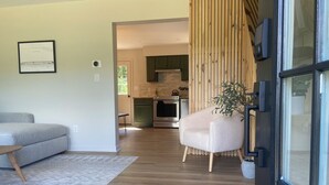 A modern living room with a grey sofa, a white armchair, and a wooden floor. There is a kitchen in the background with dark cabinets and stainless steel appliances. - Serene Mid-Mod Ranch on 2 Acres in Gardiner (Gardiner)