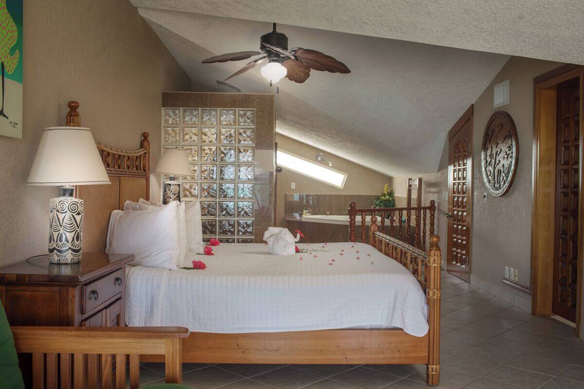 A bedroom with a bed, wooden headboard, bedside table with a lamp, a ceiling fan, and a decorative wall panel.