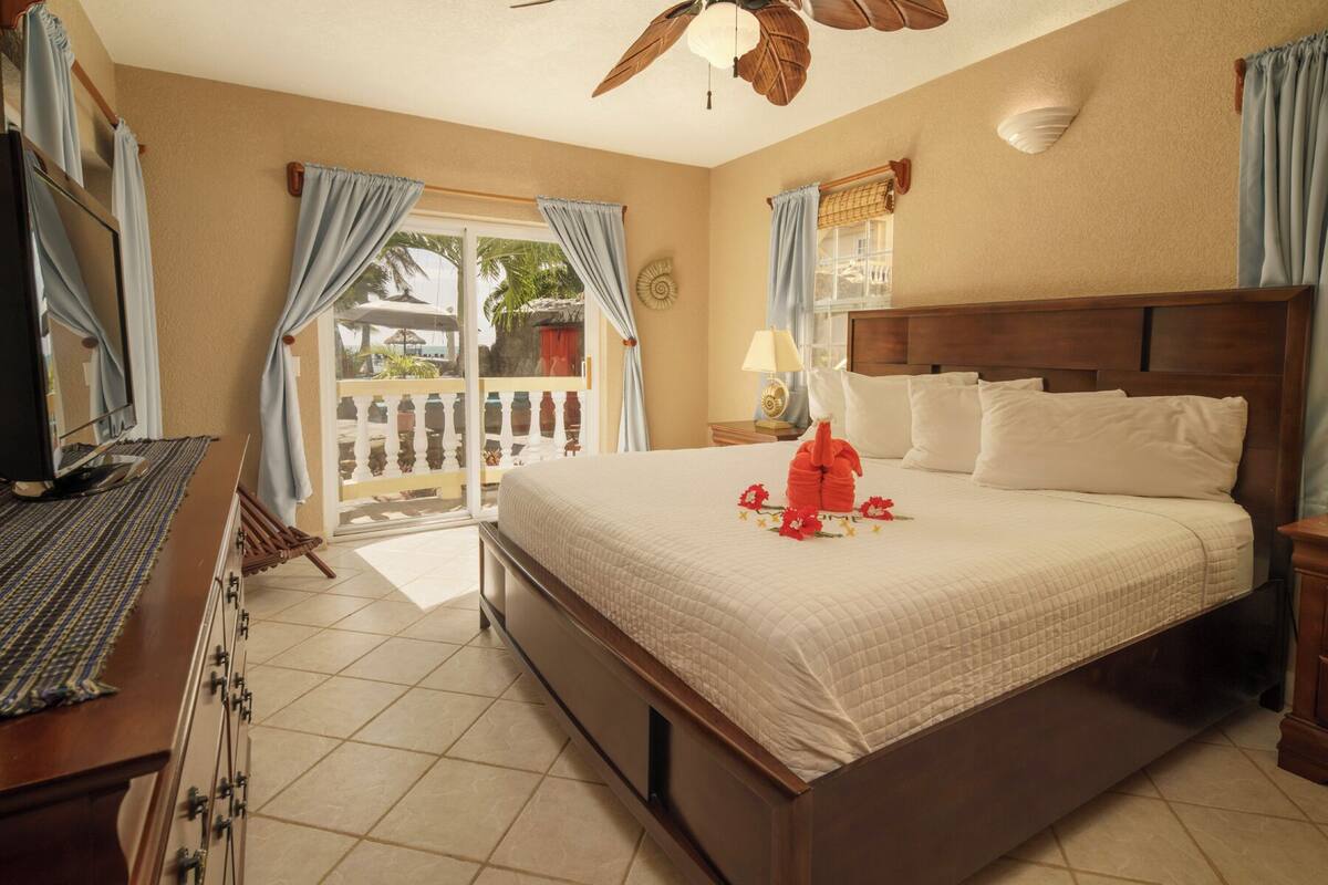 A hotel room with a bed, a desk with a computer, a window with curtains, and a balcony with a view of palm trees.