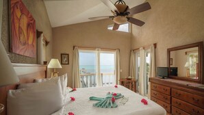 Habitación de hotel con cama, ventilador de techo, televisor, cómoda y un gran ventanal con vista al océano.