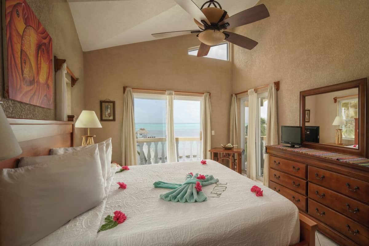 A hotel room with a bed, a ceiling fan, a TV, a dresser, and a large window with a view of the ocean.