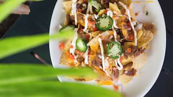 Un plato de nachos con ingredientes adicionales, sobre una superficie oscura con follaje verde al frente.