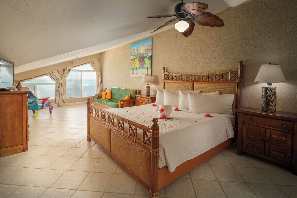 A spacious bedroom with a large bed, a ceiling fan, and a view of the sea through a window.