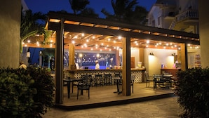 Zona de bar al aire libre con mesas y sillas de madera, luces colgantes y vistas de la ciudad al atardecer.