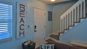 A hallway with a staircase, a door, a wall-mounted sign reading "BEACH", and a cushion with the text "LIFE IS BETTER". - Flip Flop Living 1 3bdrm, 2.5 bath, STEPS TO SAND! (New Smyrna Beach)