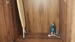 Un meuble en bois doté d’une planche à repasser intégrée et d’un petit appareil électroménager, vraisemblablement un fer à vapeur, posé sur une étagère.