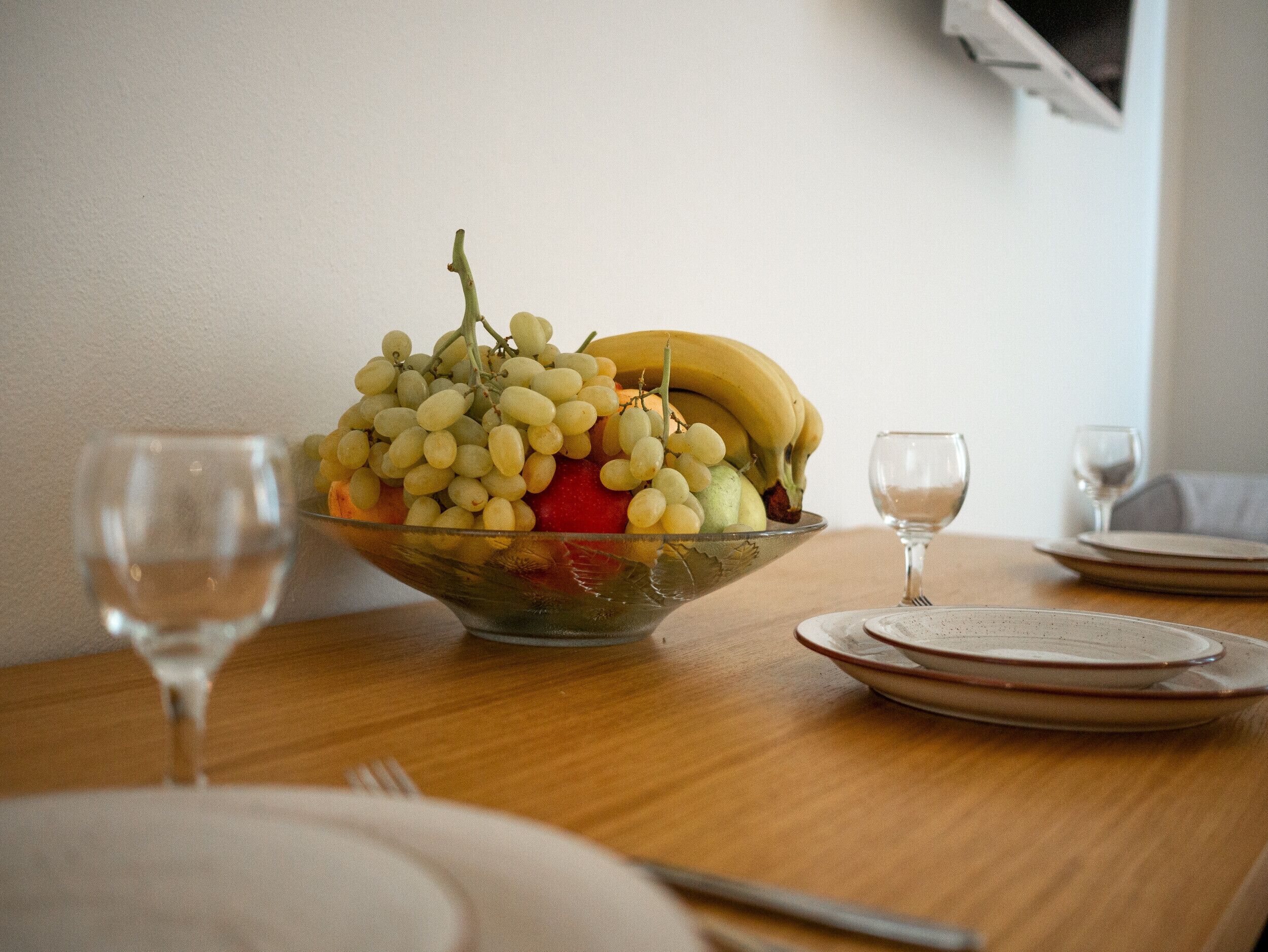 A dining table with a fruit bowl containing grapes, bananas, and strawberries. There are wine glasses and plates on the table.