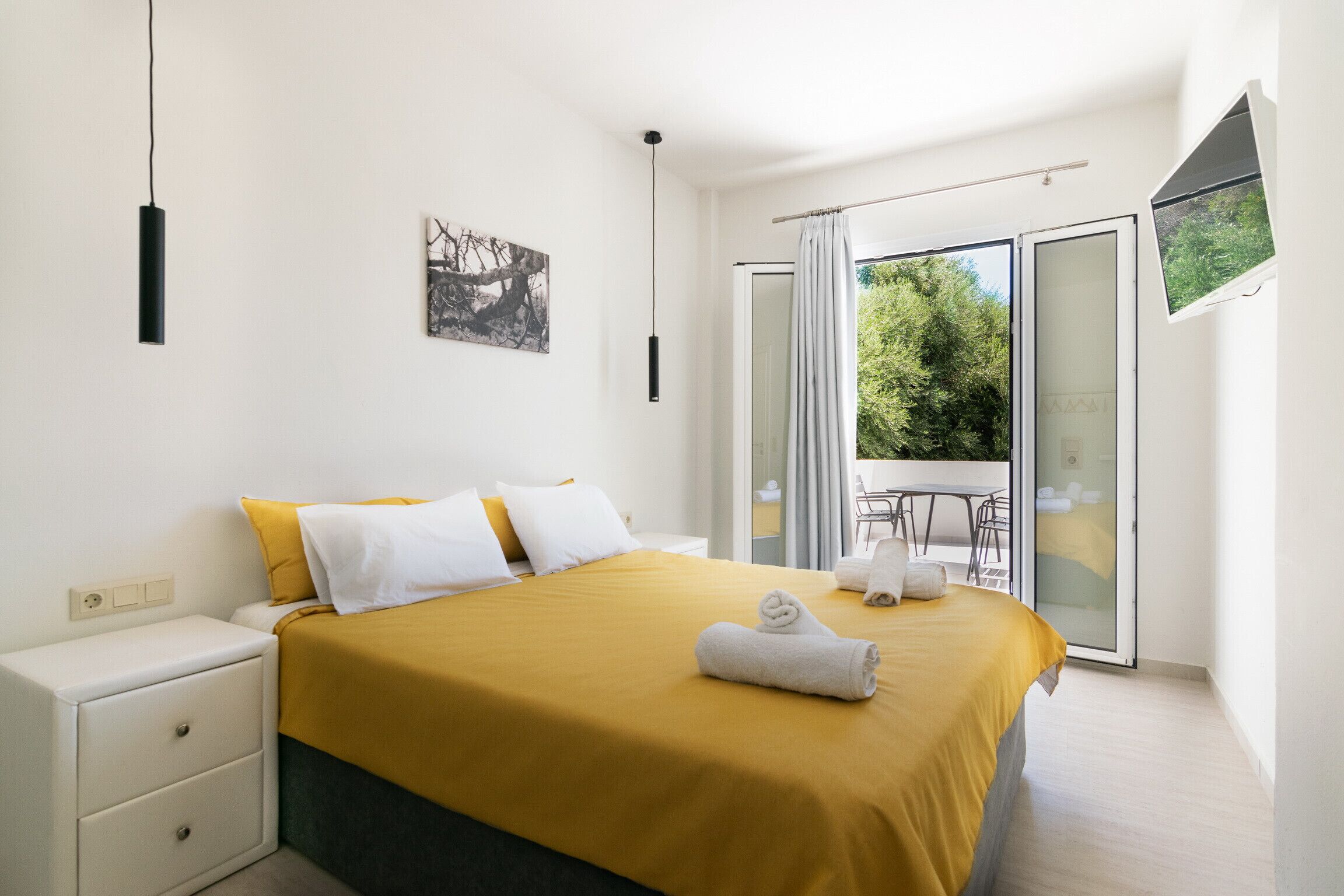 A modern bedroom with a large bed, a nightstand, a painting, and a balcony with a table and chairs.