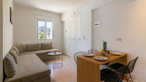 A modern living room with a sofa, a dining table with chairs, a small round table, and a window with a view of the outdoors. - Secretnest 15, apartment near the beach,Plakias (Plakias)