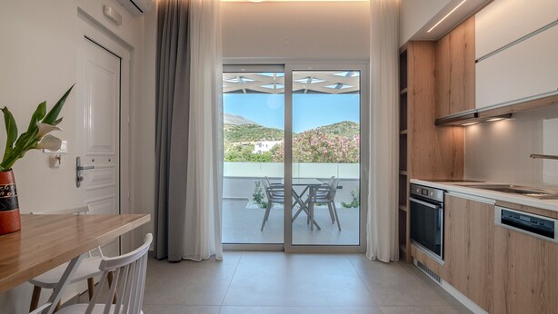 A modern kitchen with wooden cabinets, a built-in oven, and a dishwasher. There is a dining area with a table and chairs, and a sliding glass door leading to a balcony with a view of greenery and hills.