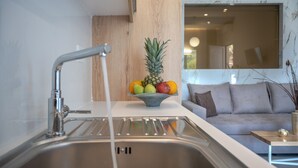 A modern kitchen with a stainless steel sink, a bowl of fruit on the countertop, and a grey sofa in the background.