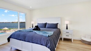 Un dormitorio con un ventanal grande, una cama con cabecero blanco, dos mesitas de noche y vista al mar.