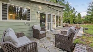 A patio with wicker furniture and a stone floor, adjacent to a house with large windows and a covered entryway. - Mid-Century Nest, Near Forest Park (Portland)