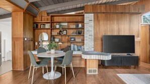 A modern living room with a wooden bookshelf, a round table with chairs, a flat-screen TV, and a stone accent wall. - Mid-Century Nest, Near Forest Park (Portland)