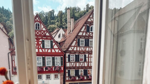 Vista a través de una ventana de casas tradicionales con entramado de madera, jardineras con flores y un mapa en el alféizar.