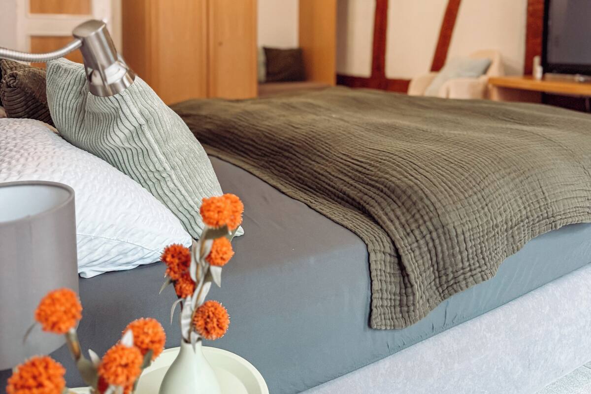 Un dormitorio con cama, almohadas, mesita de noche, lámpara y un jarrón con flores.