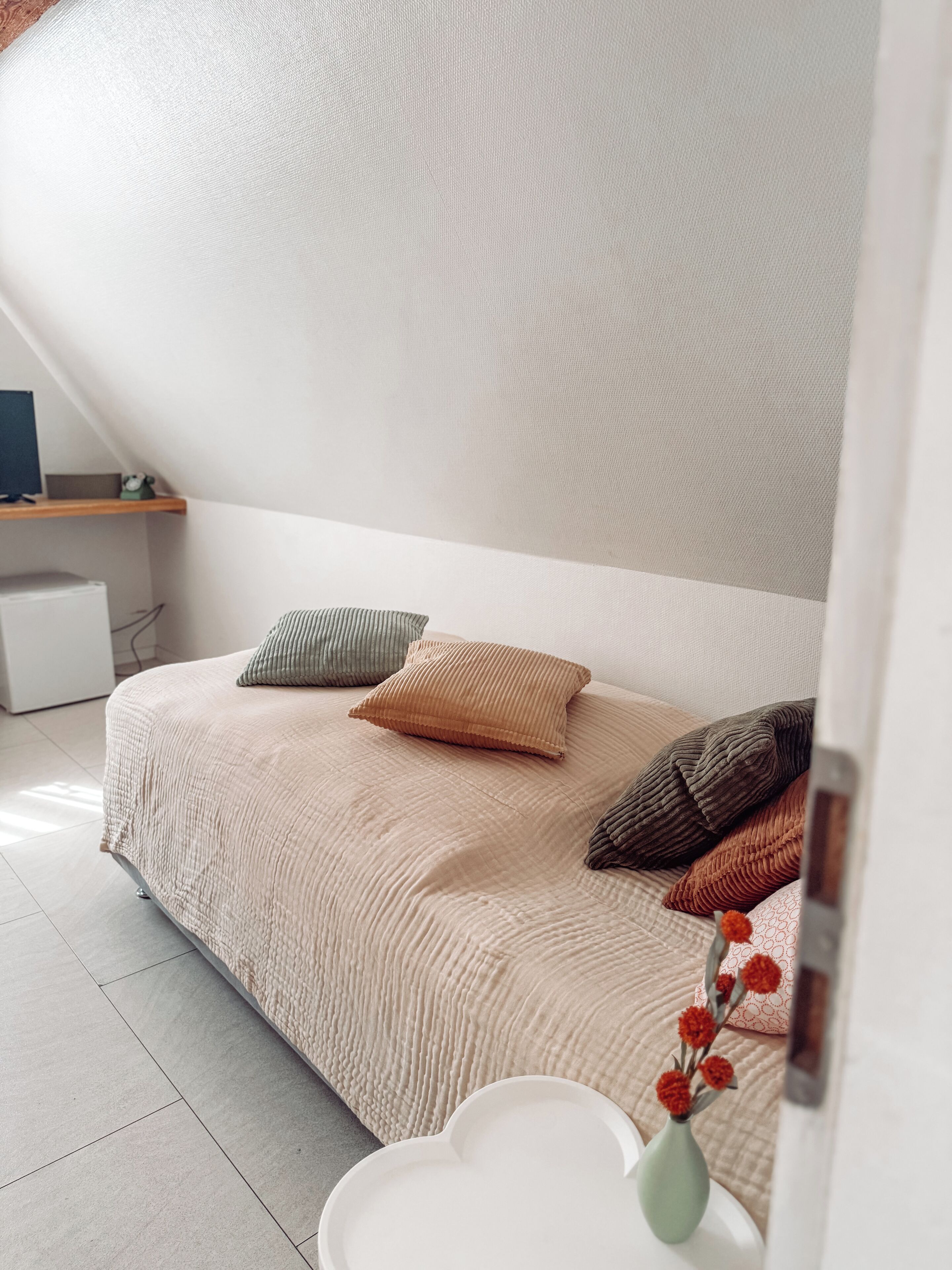 Un dormitorio con una cama, almohadas, una mesita de noche con un jarrón y un escritorio con un ordenador.