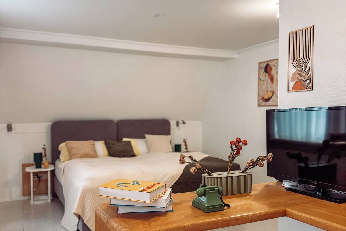 Una habitación de hotel moderna con una cama bien hecha, una mesita de noche de madera con libros y un teléfono, un televisor de pantalla plana y un jarrón decorativo con flores.
