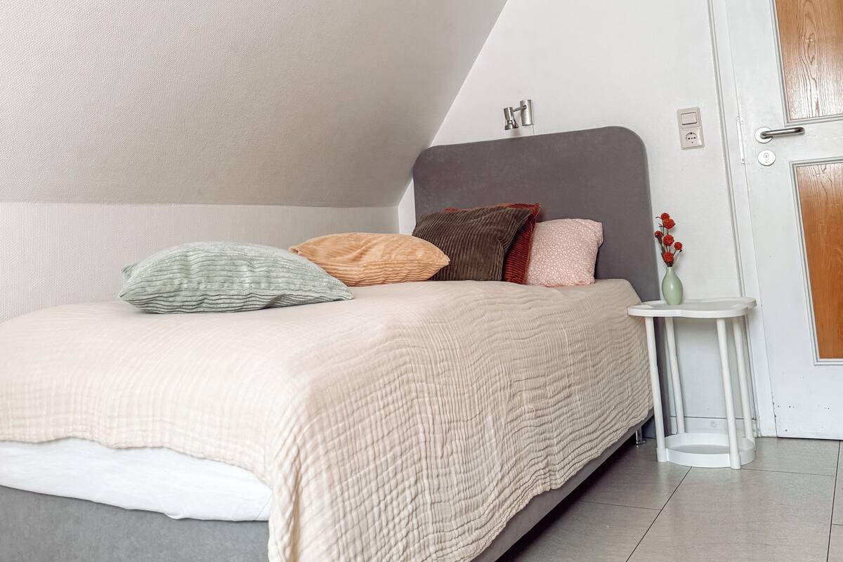 Un dormitorio con techo inclinado, una cama con muchas almohadas, una mesita de noche con un florero y una puerta con panel de madera.