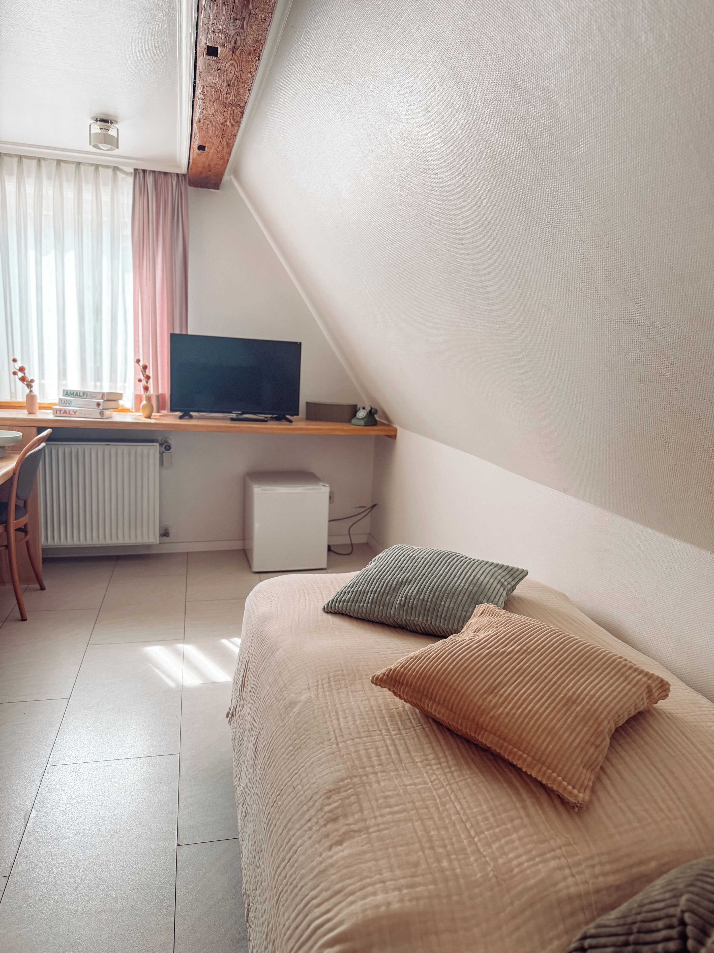 A bedroom with a bed, a desk with a computer, a radiator, and a window with curtains.