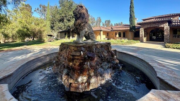 Una fuente de piedra con una estatua de león en una zona ajardinada, con un edificio al fondo.