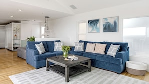 A modern living room with a sectional sofa, a coffee table, and a rug. There are two paintings on the wall and a plant on the side table.