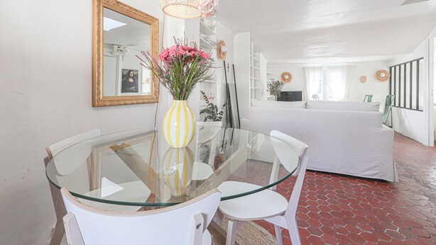 A dining area with a glass-top table, white chairs, a vase with flowers, a mirror, and a white sofa in the background.