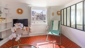 A room with a TV, a window with curtains, a green chair, and a view of buildings.