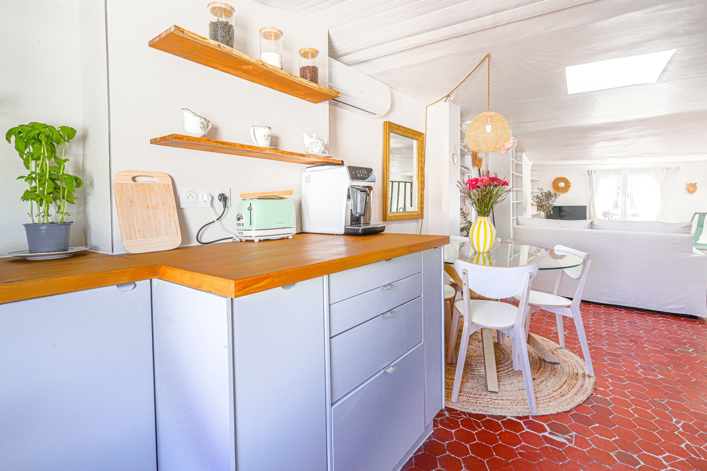 Cozinha compacta com bancada de madeira, geladeira embutida, cafeteira e torradeira. Há uma área de refeições com mesa e cadeiras, e um vaso com planta.