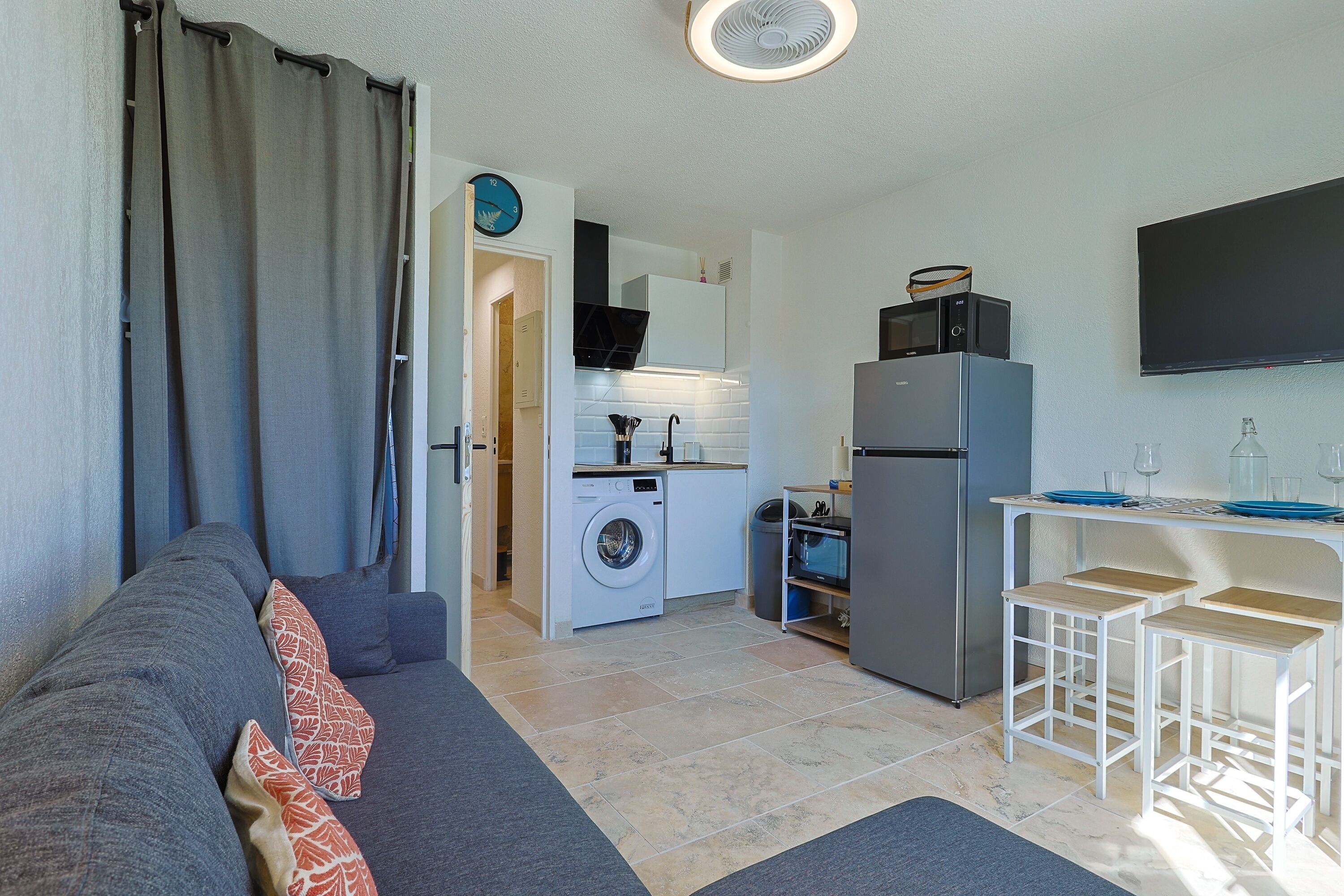 A compact living space with a sofa, a washing machine, a refrigerator, and a TV mounted on the wall.