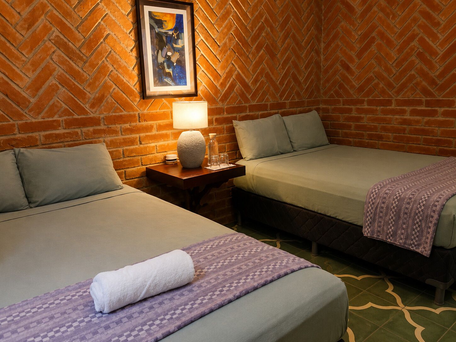 A hotel room with two beds, a brick wall, a framed picture, a nightstand with a lamp, and a patterned rug.