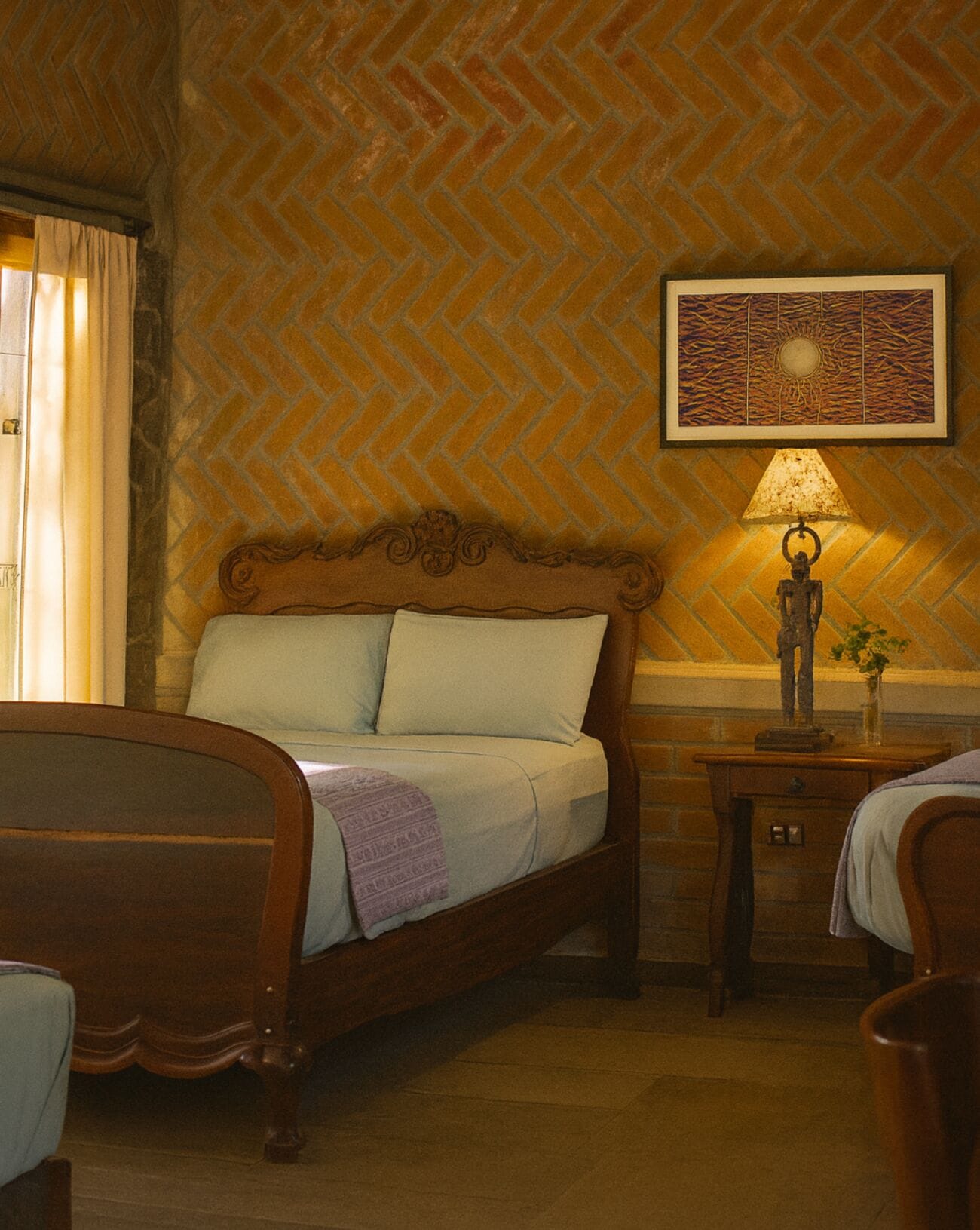 Une chambre avec un mur en briques disposées en motif chevrons, un lit doté d’une tête de lit en bois, une table de chevet avec une lampe, et une œuvre d’art encadrée au-dessus du lit.