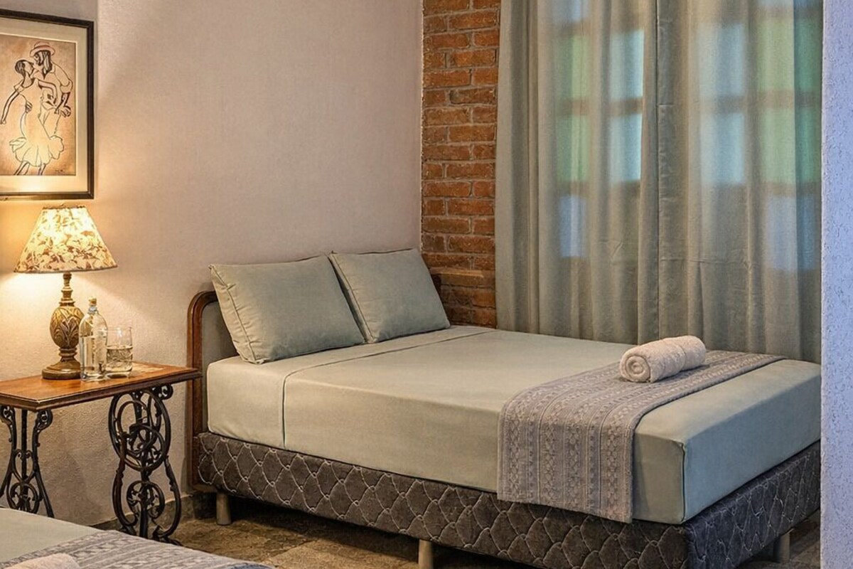 Una camera da letto con un letto, un comodino, una lampada, una finestra con tende e un muro in mattoni.