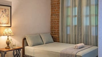 Um quarto com uma cama, mesa de cabeceira, luminária, janela com cortinas e uma parede de tijolos.
