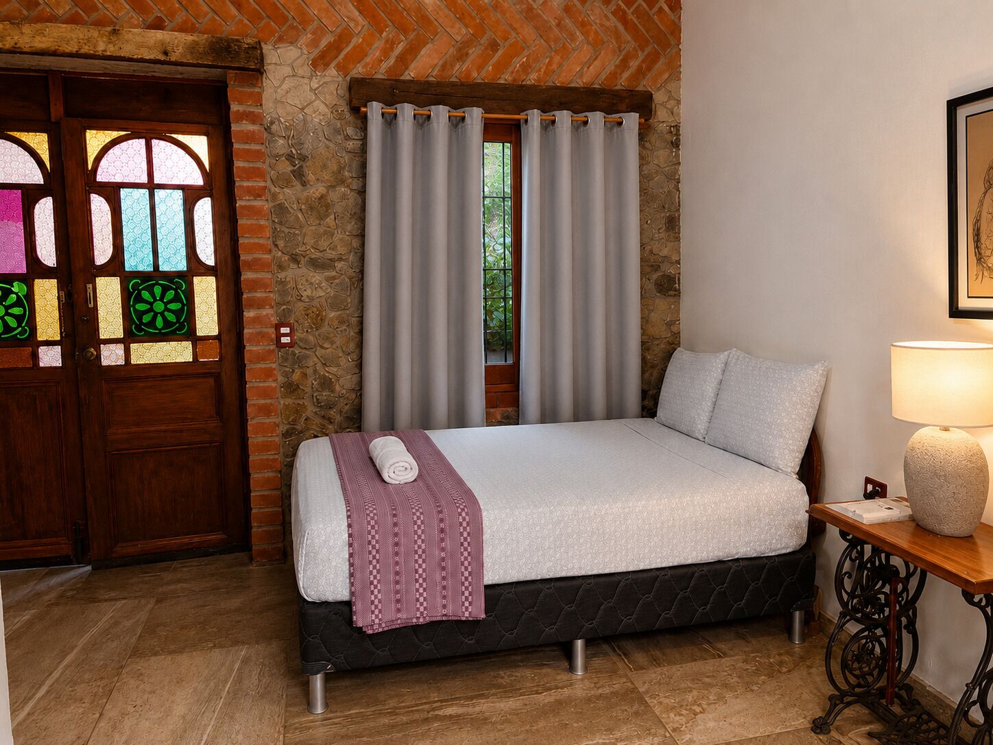 Un dormitorio con una cama, una ventana con cortinas, una puerta de madera con vitrales y una mesita de noche con una lámpara.