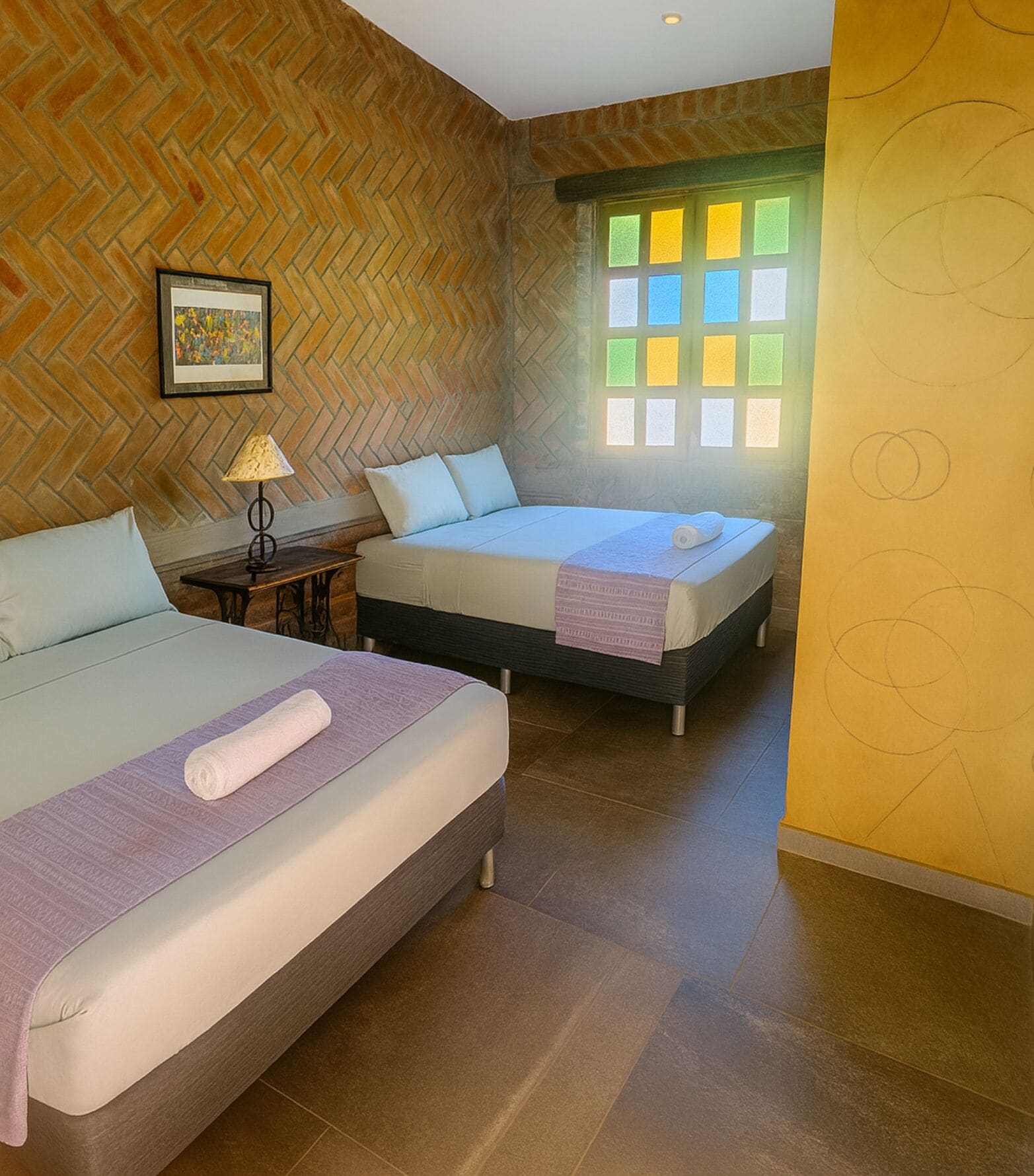 Una habitación de hotel con dos camas, un muro de ladrillo con patrón de espiga, un ventanal con vidrios de colores y un cuadro enmarcado en la pared.