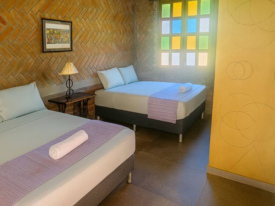 Una habitación de hotel con dos camas, un muro de ladrillo con patrón de espiga, un ventanal con vidrios de colores y un cuadro enmarcado en la pared.