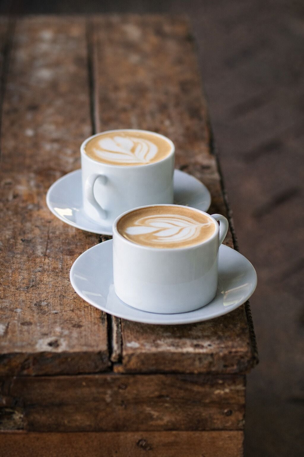 Dos tazas blancas de café con diseños de espuma sobre una mesa de madera rústica.