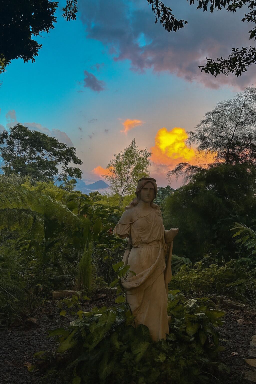 Una estatua en un jardín frondoso con un atardecer de fondo.