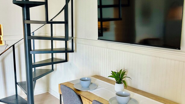 A modern dining area with a wooden table, two grey chairs, a small potted plant, and a black spiral staircase. - 2 bedroom Modern Retreat (Baton Rouge)