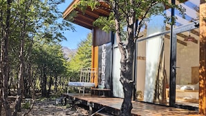 Una terraza de madera con un banco, un vidrio y un árbol frente a un edificio.