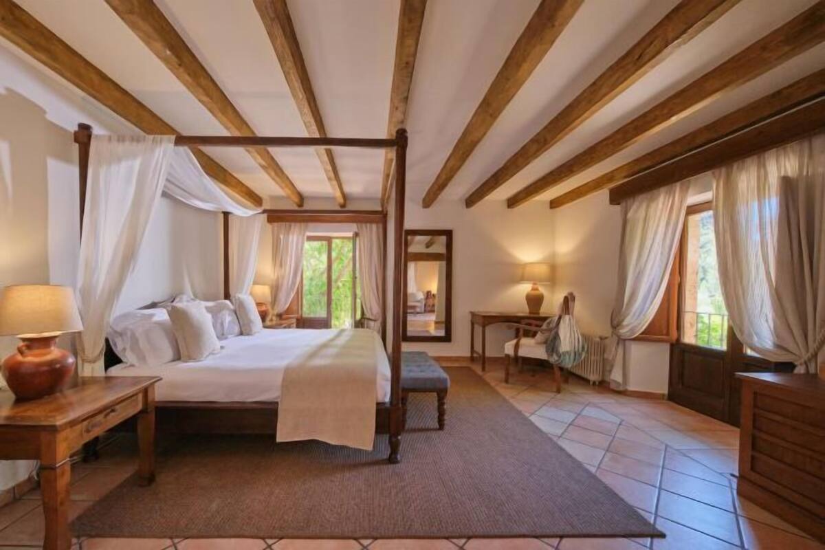 Ein Schlafzimmer mit einem Himmelbett, einem Holztisch, einem Stuhl und einem Fenster mit Vorhängen.