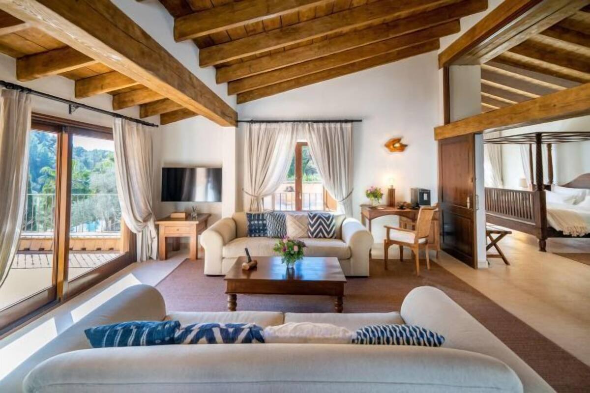 Ein geräumiger Wohnbereich mit Sofa, Couchtisch und Fernseher. Es gibt Fenster mit Vorhängen, einen Balkon und ein Bett mit Holz-Kopfstück.