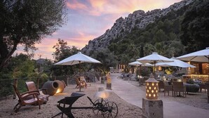 Un área de estar al aire libre con sombrillas, sillas y una chimenea, rodeada de árboles y montañas al fondo.