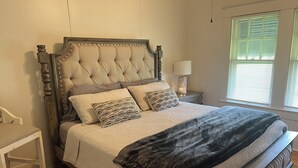 A bedroom with a tufted headboard, a bed with patterned pillows, a bedside table, and a window with blinds. - Quiet, Peaceful River property on 1.5 acres in Sterlington. (Sterlington)